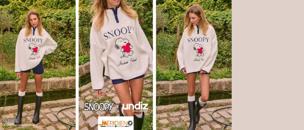 Femme portant un pull polaire blanc à col bleu marine avec l’inscription « Snoopy Social Club » et un motif de Snoopy tenant un cœur rouge. Elle se promène dans un jardin verdoyant, habillée d’un short bleu et de bottes noires, dans une ambiance automnale et naturelle.